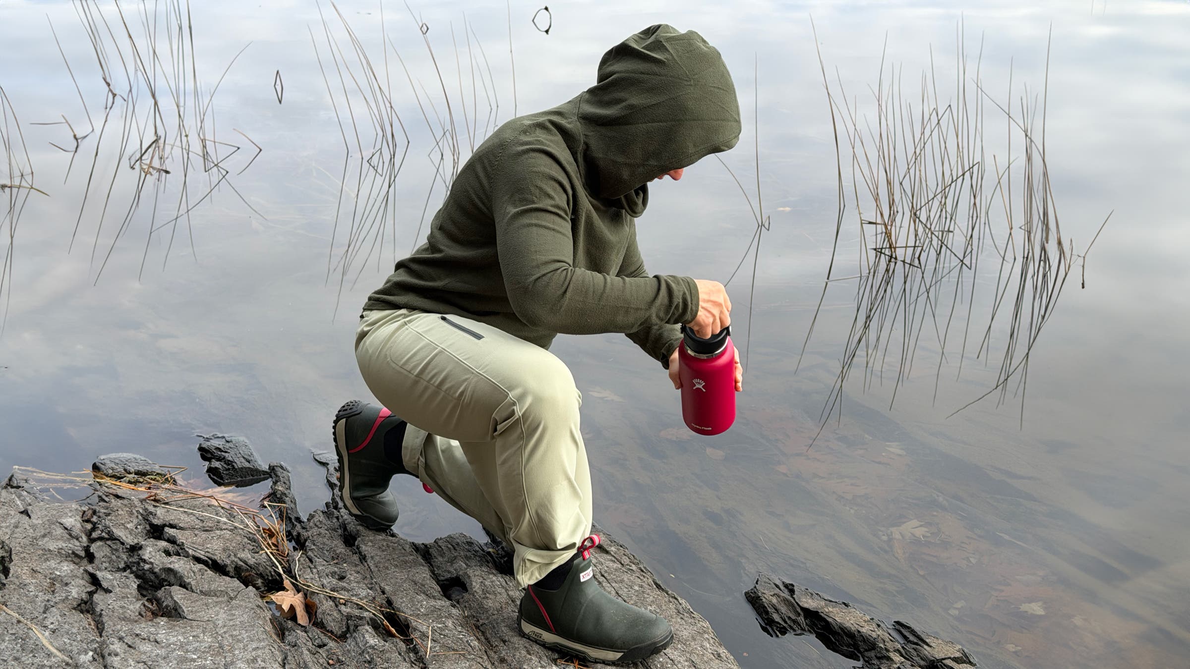 Hiker filling up water bottle on shoreline of lake wearing flexible and lightweight Stio Pinedale Pant.