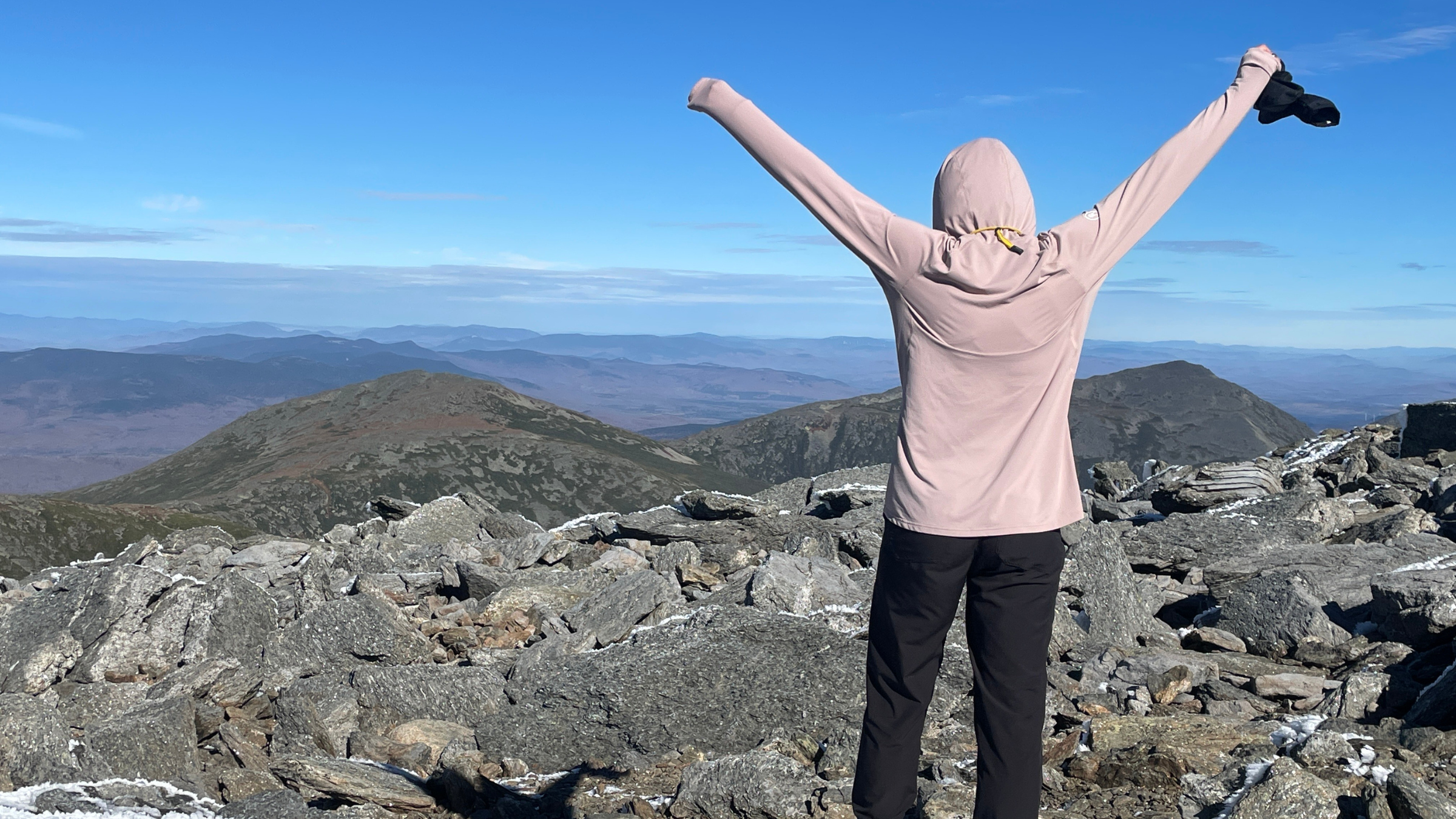 woman stands with back to camera wearing a women's best sun hoodie from Backcountry, arms raised at top of mountain looking at view