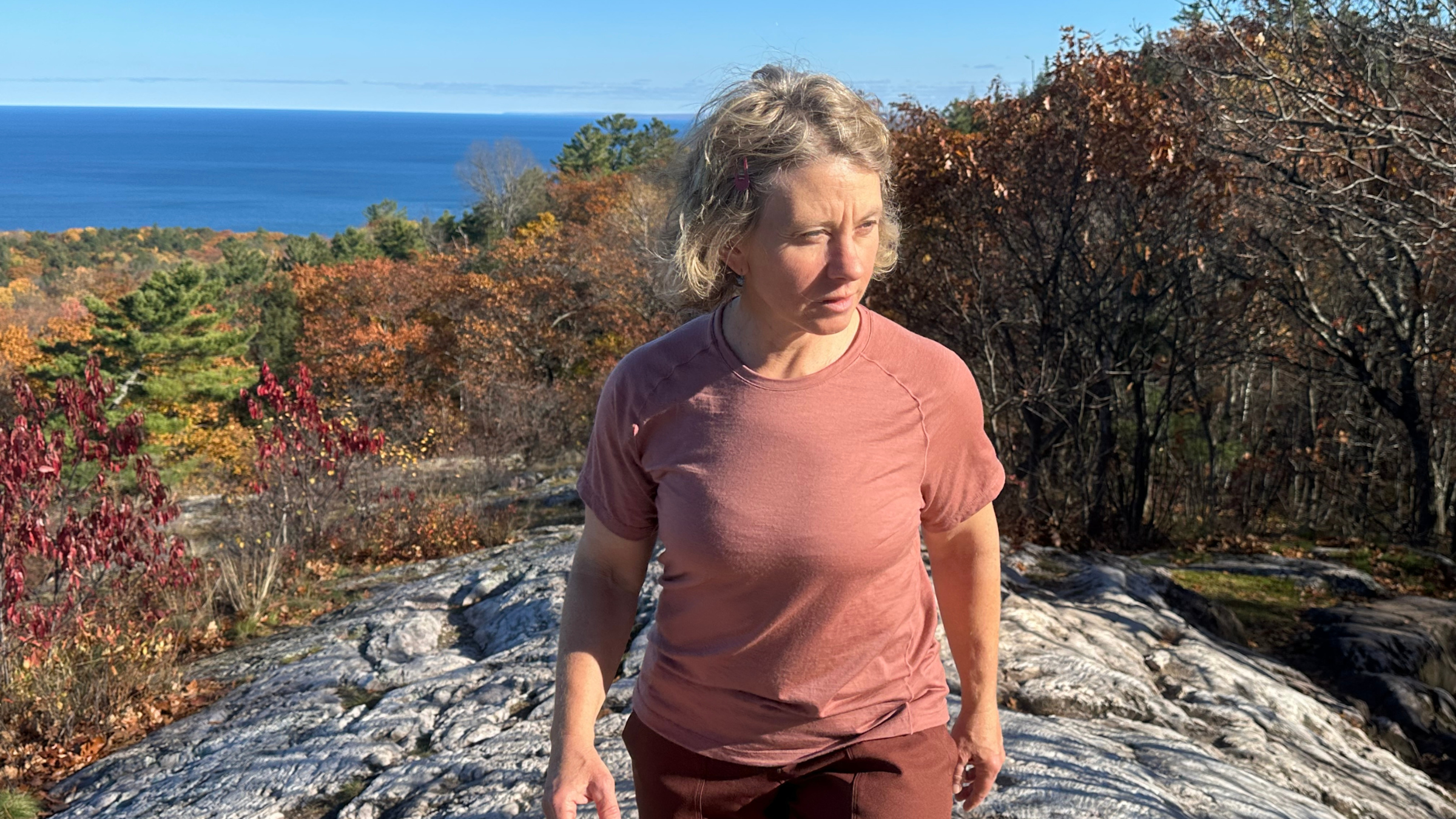 woman walking toward camera in dark pink best women's hiking tee with rocks, trees in fall colors and water in the background