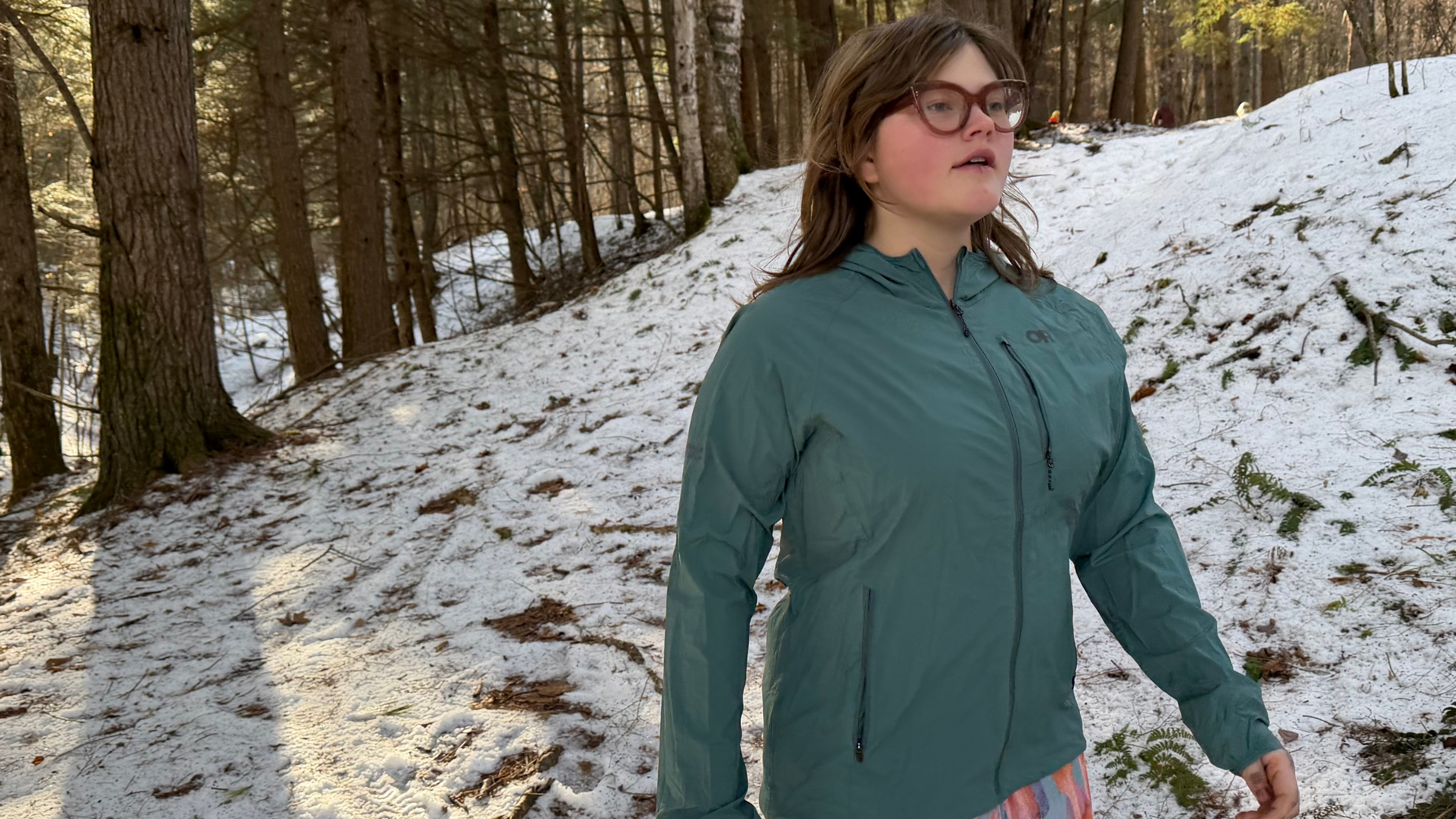 young woman in glasses walking through forest with snow on ground wearing green Outdoor Research best windshell, best hiking shirts for women